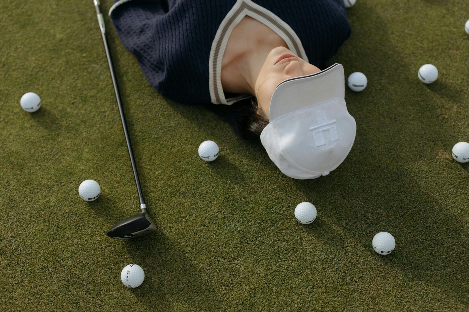 A high-angle, close-up shot of a person lying flat on their back on a lush green golf course. They are wearing a dark navy ribbed sweater with a striped V-neck and a white baseball cap pulled down over their eyes. Several white golf balls are scattered around them on the grass, and a golf club lies positioned to the left.